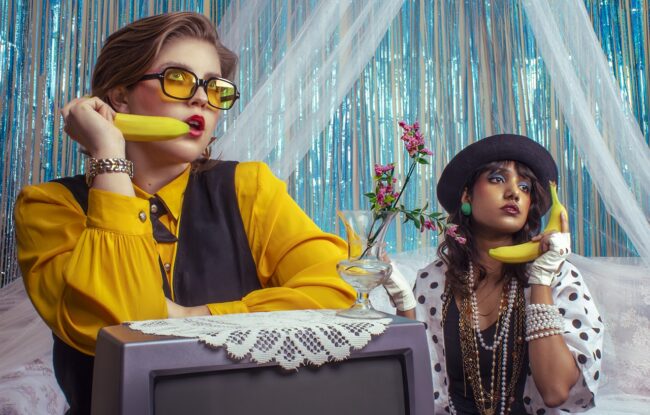 Vanessa, a feminine-presenting woman in a vibrant yellow outfit and matching glasses, sits to the right of the frame, playfully holding a banana like a phone. On the left, Ashanti, also a feminine-presenting woman, mirrors her gesture, holding a banana as if making a call.