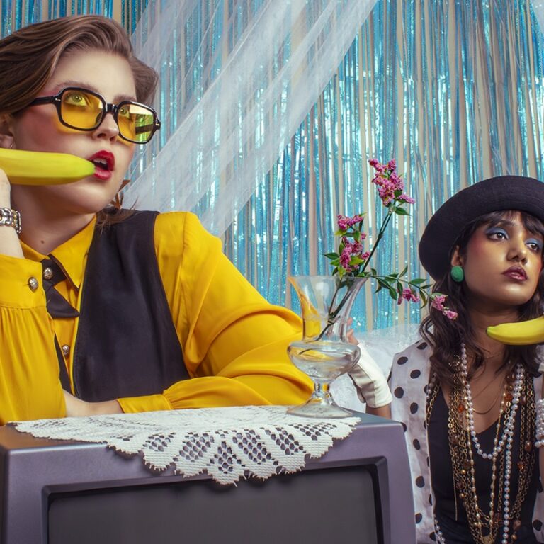 Vanessa, a feminine-presenting woman in a vibrant yellow outfit and matching glasses, sits to the right of the frame, playfully holding a banana like a phone. On the left, Ashanti, also a feminine-presenting woman, mirrors her gesture, holding a banana as if making a call.