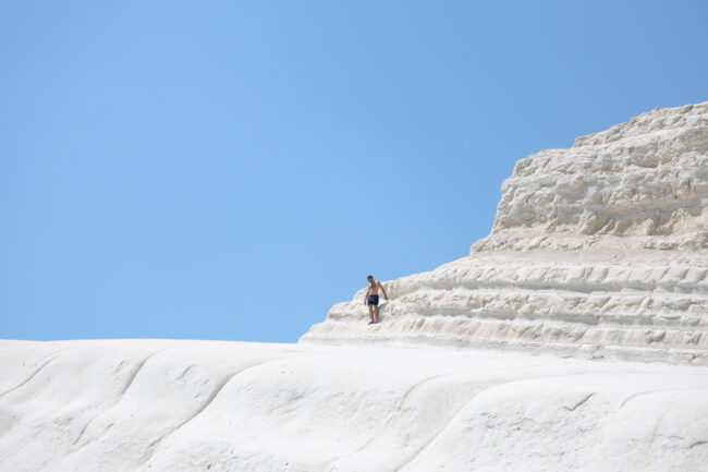 A lone figure is captured standing in the centre of a large white rocky cliff with a pale blue sky appearing in the background.