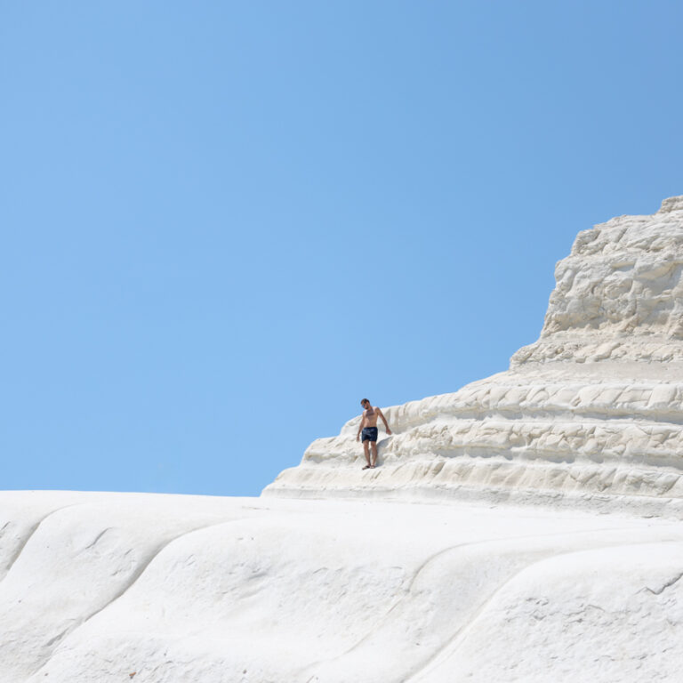 A lone figure is captured standing in the centre of a large white rocky cliff with a pale blue sky appearing in the background.