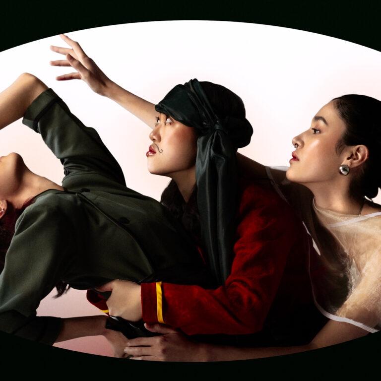 Image of 3 women wearing moustache makeup and asia traditional clothes. Model 1 (first from left to right), is looking up. The other models are looking in the same direction towards the left. Bright red-gradient background. Dark green, sharp oval frame is overlayed on top of the composition.