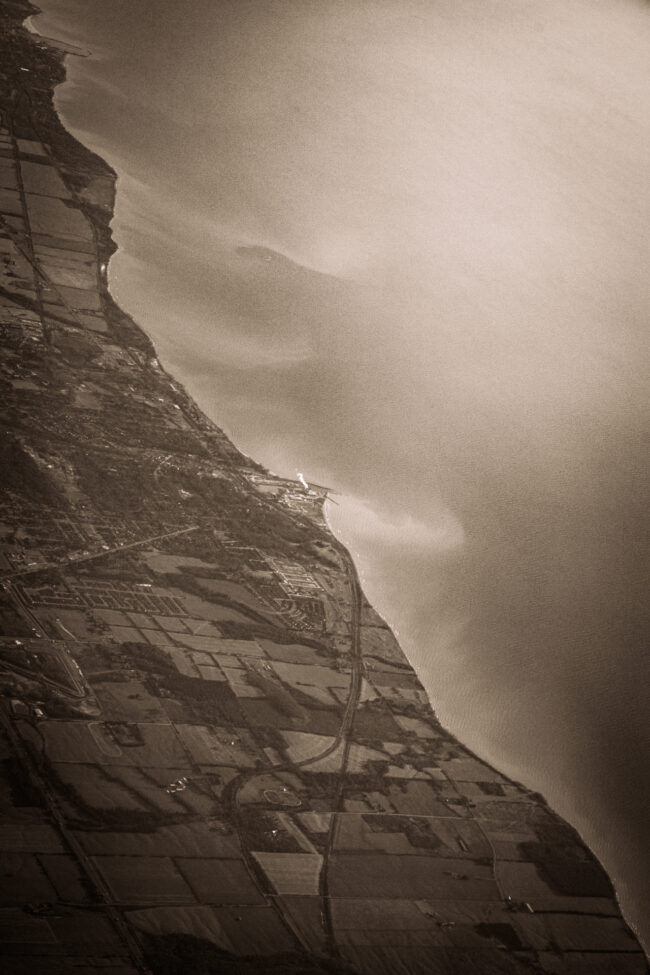 The image is composed of two parts divided diagonally from top left to bottom right. The left half is composed of the land, and farms. The roads on the land all lead towards the centre of the image, which has a very proportionately small factory that is letting of a smoke cloud that dwarfs the factory itself. The right half is made up of a lake. The water on the lake seems to have something from the land bleeding into it as tendrils of sand, dozens of meters long, reach out into the lake.