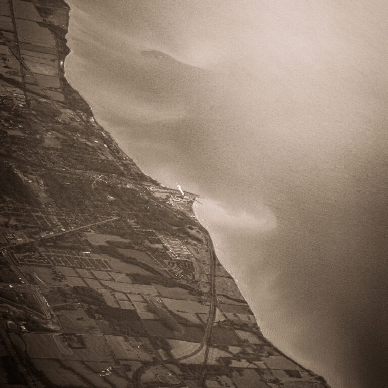 The image is composed of two parts divided diagonally from top left to bottom right. The left half is composed of the land, and farms. The roads on the land all lead towards the centre of the image, which has a very proportionately small factory that is letting of a smoke cloud that dwarfs the factory itself. The right half is made up of a lake. The water on the lake seems to have something from the land bleeding into it as tendrils of sand, dozens of meters long, reach out into the lake.