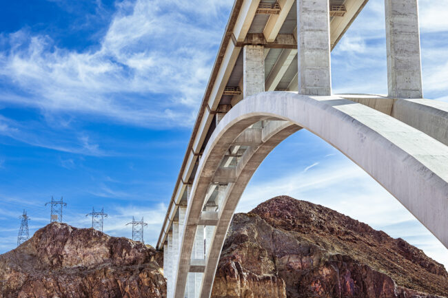 A photo of a large bridge, reaching over to a rocky landmass. On the left of the landmass sits four power line poles. the sky is blue and partly cloudy with cirrus clouds.