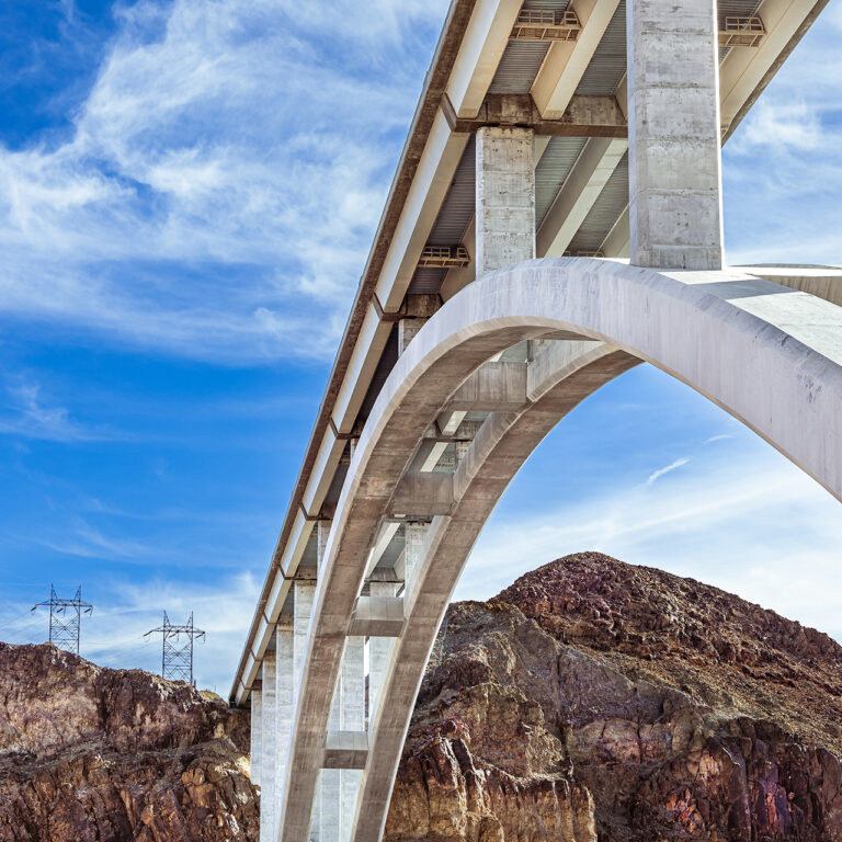 A photo of a large bridge, reaching over to a rocky landmass. On the left of the landmass sits four power line poles. the sky is blue and partly cloudy with cirrus clouds.