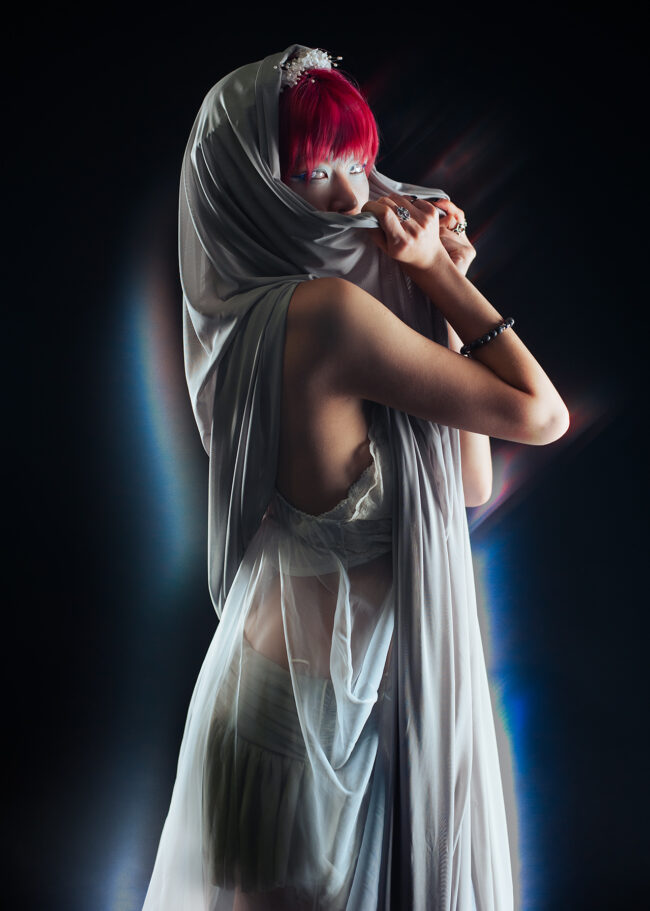A woman in bright red hair, hooded in a grey fabric. She looks at the camera mysteriously, using the fabric to cover the bottom part of her face. Light refractions surround her, giving a glowing effect.