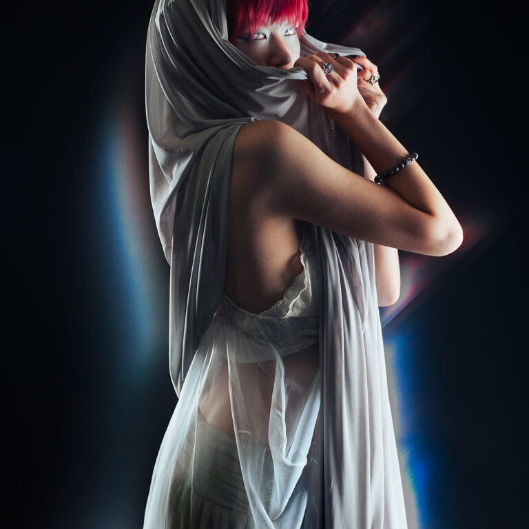 A woman in bright red hair, hooded in a grey fabric. She looks at the camera mysteriously, using the fabric to cover the bottom part of her face. Light refractions surround her, giving a glowing effect.
