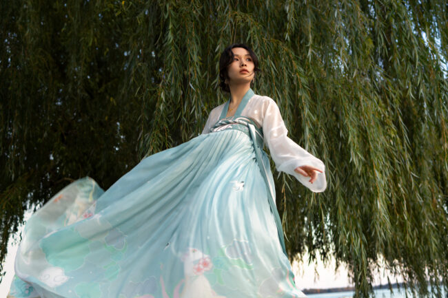 The image features a Chinese model dressed in a light teal Hanfu, positioned at the center of the frame. The long silk dress flows gracefully as the model sways, showcasing its delicate pattern and movement. A soft orange glow from the setting sun illuminates the model's face, creating a serene, warm atmosphere. The model stands under a tree with long leaves, their green and orange hues softly framing the scene and blending with the overall natural palette. The image captures a moment of quiet reflection, blending the elegance of the Hanfu with the beauty of autumn.