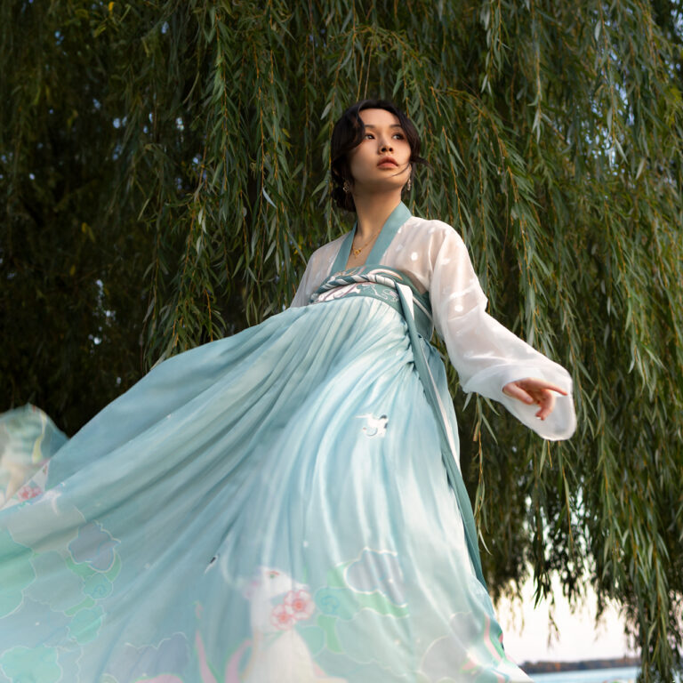 The image features a Chinese model dressed in a light teal Hanfu, positioned at the center of the frame. The long silk dress flows gracefully as the model sways, showcasing its delicate pattern and movement. A soft orange glow from the setting sun illuminates the model's face, creating a serene, warm atmosphere. The model stands under a tree with long leaves, their green and orange hues softly framing the scene and blending with the overall natural palette. The image captures a moment of quiet reflection, blending the elegance of the Hanfu with the beauty of autumn.