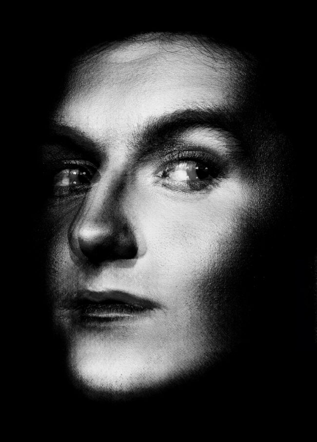 A black and white multiple exposure photograph. The image is a male presenting model at a 3/4 perspective, cropped to show just the face emerging from the darkness.