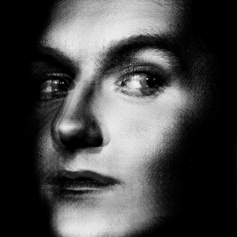 A black and white multiple exposure photograph. The image is a male presenting model at a 3/4 perspective, cropped to show just the face emerging from the darkness.