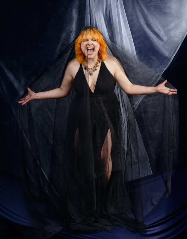 A young woman with bright orange hair in a shag cut stands in front of a layered, fabric backdrop. She is covered in a fine veil, and has her arms outstretched. She is slightly hunched forward into a full-chested scream. Her eyes are closed, and the scream seems to tear through her. She is in a black dress with a mid-length v-neck that is sleeveless. The backdrop behind her and the floor beneath her are both deep navy fabric, although the backdrop has tule placed in front of it, that is folding in the centre and catching light from the rear.