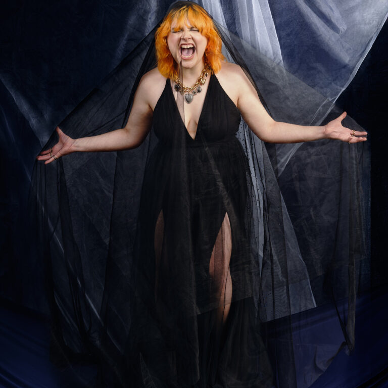 A young woman with bright orange hair in a shag cut stands in front of a layered, fabric backdrop. She is covered in a fine veil, and has her arms outstretched. She is slightly hunched forward into a full-chested scream. Her eyes are closed, and the scream seems to tear through her. She is in a black dress with a mid-length v-neck that is sleeveless. The backdrop behind her and the floor beneath her are both deep navy fabric, although the backdrop has tule placed in front of it, that is folding in the centre and catching light from the rear.