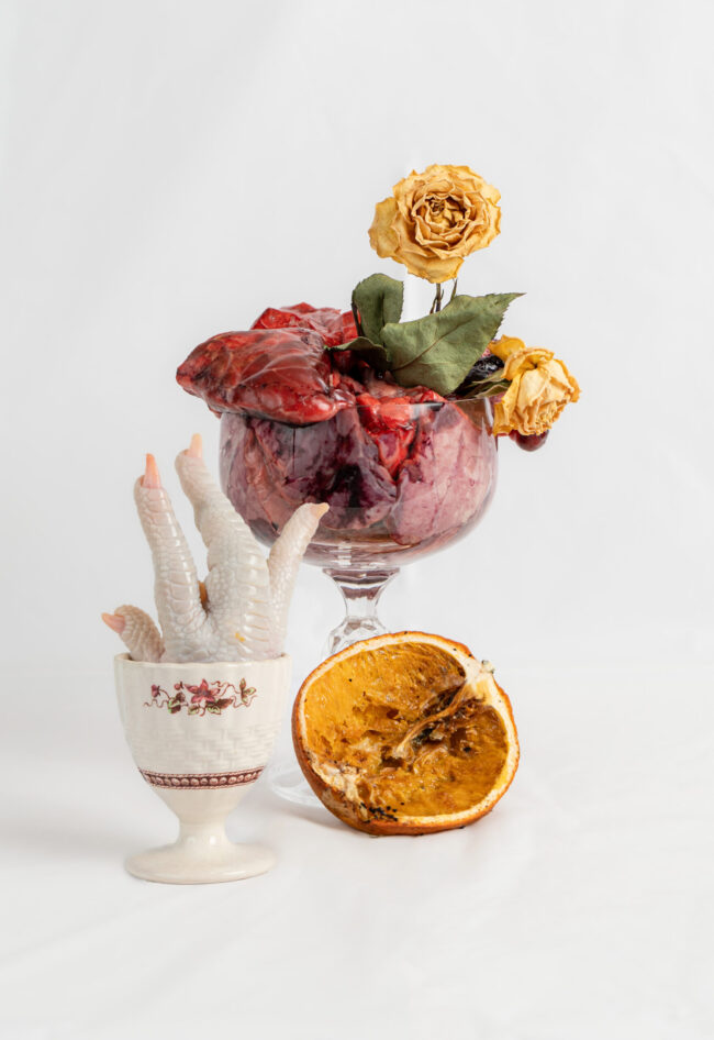 Image of chicken foot in egg stand with rotten orange and cup filled with cow lung and dying rose