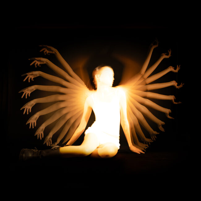A long-exposure photograph of a seated figure with multiple arms radiating outward, resembling wings, with warm golden light illuminating the motion.