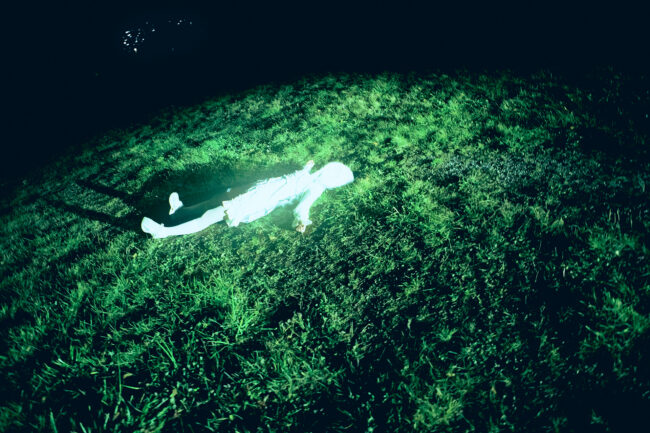 An overhead shot of a faceless figure dressed entirely in white, lying down spread-eagled in a field of green grass. Shadows engulf the edge of the frame, with the grassy field fading out into darkness.