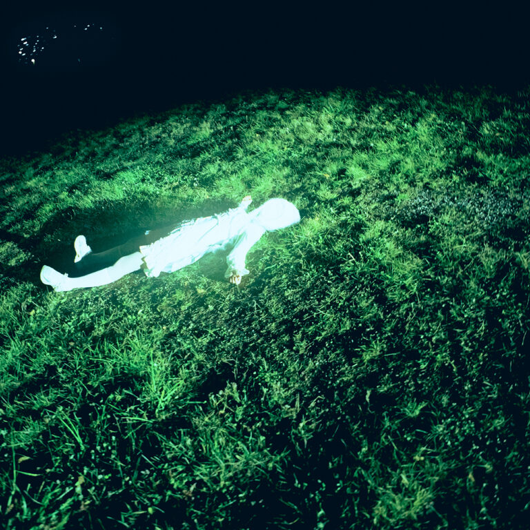An overhead shot of a faceless figure dressed entirely in white, lying down spread-eagled in a field of green grass. Shadows engulf the edge of the frame, with the grassy field fading out into darkness.