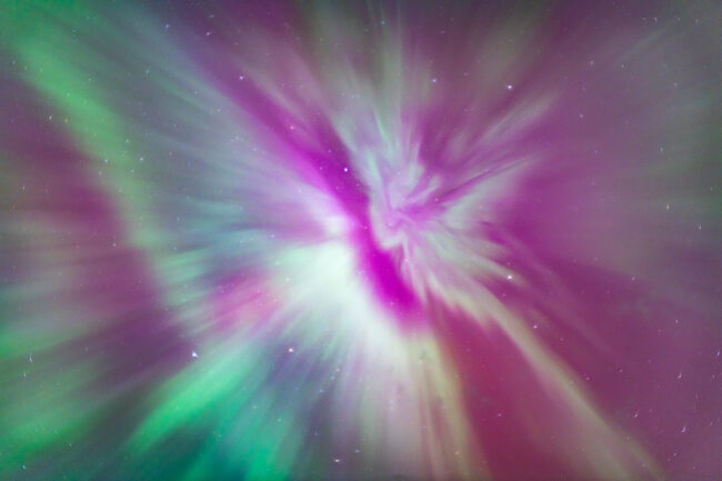 Worm's-eye view of an aurora corona captured during an aurora storm. Bright, saturated green and pink bands of the aurora borealis morph and bend across the starry night sky. They appear to create a crown-like shape as they converge toward a middle point.