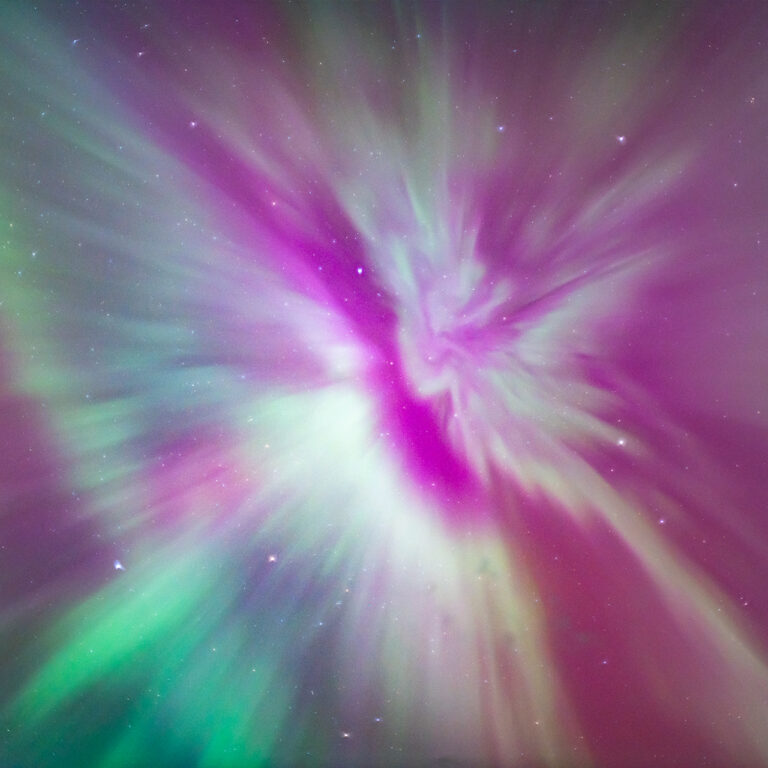 Worm's-eye view of an aurora corona captured during an aurora storm. Bright, saturated green and pink bands of the aurora borealis morph and bend across the starry night sky. They appear to create a crown-like shape as they converge toward a middle point.