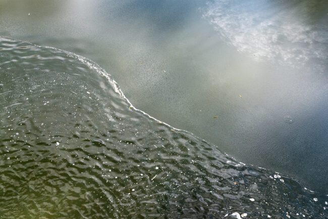 A landscape composition of a half-frozen pond. Water washes over the ice, creating a waving diagonal split between the bottom and top halves of the scene. Mossy green and cerulean blue tones are present throughout. White patterns in the ice at the top right corner of the frame appear almost cloud-like.