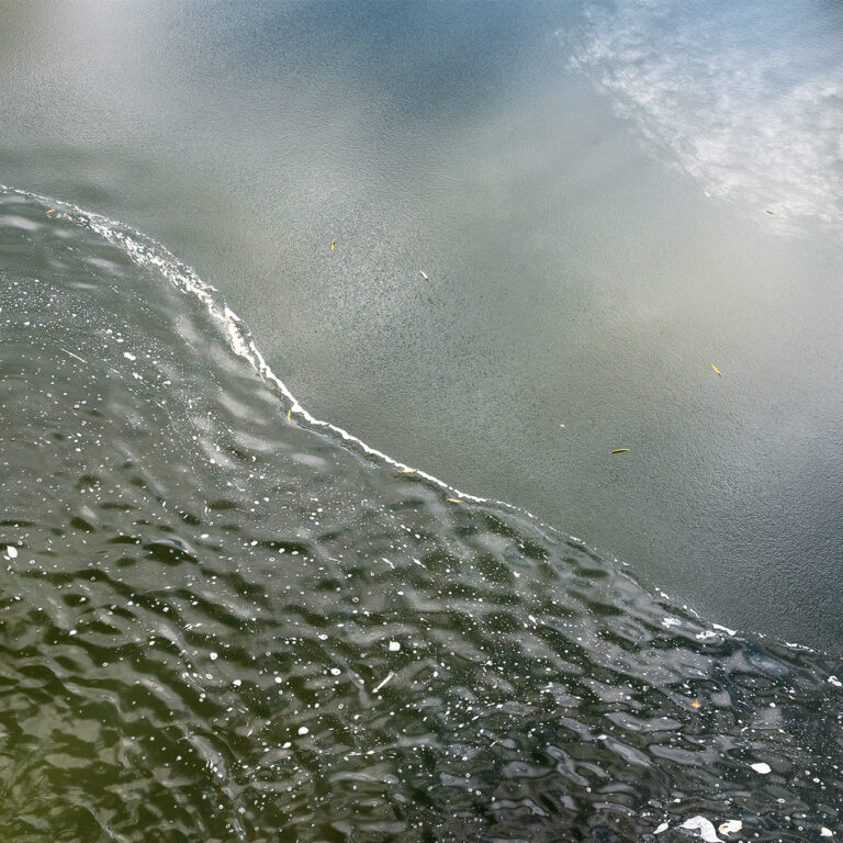 A landscape composition of a half-frozen pond. Water washes over the ice, creating a waving diagonal split between the bottom and top halves of the scene. Mossy green and cerulean blue tones are present throughout. White patterns in the ice at the top right corner of the frame appear almost cloud-like.