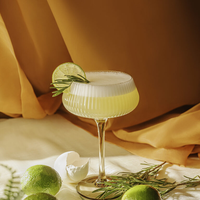 A pisco sour with a frothy top sits in an elegant glass, garnished with a lime slice and rosemary sprig. Surrounded by fresh limes, rosemary, and a cracked eggshell, the drink rests on a soft, light-colored surface. Warm golden fabric in the background casts a cozy, sunlit glow, enhancing the refined and refreshing aesthetic.