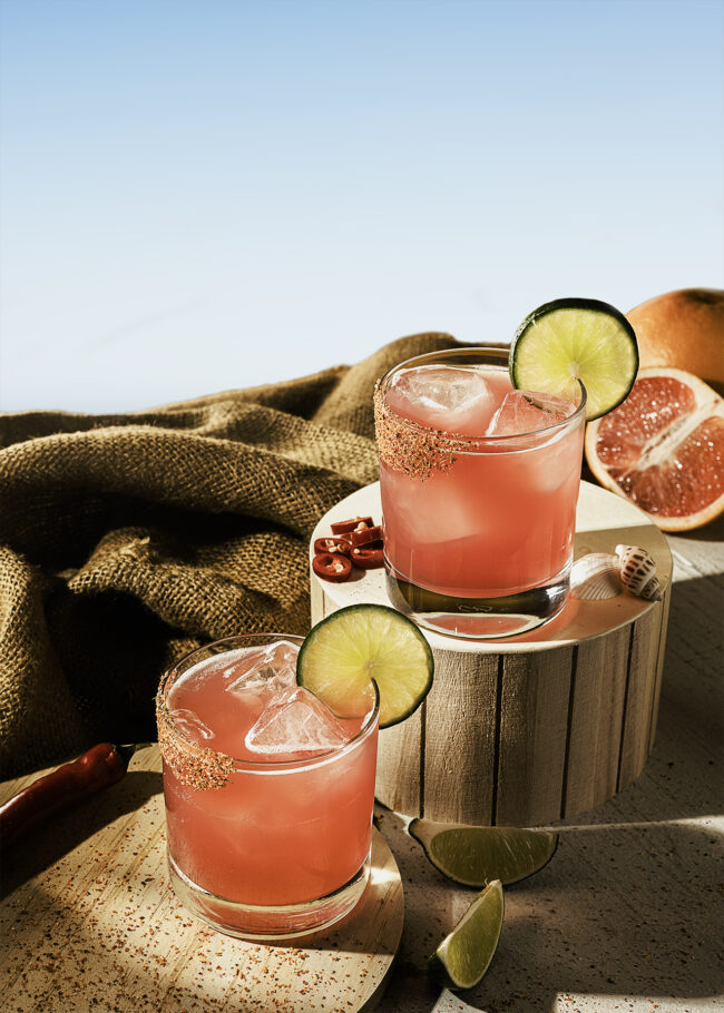 This image shows two pink margaritas in short glasses with ice, garnished with lime slices and chili-salt rims. The rustic setting includes burlap fabric, wooden pedestals, chili peppers, and grapefruit halves, creating a warm, summery vibe.