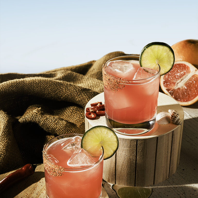 This image shows two pink margaritas in short glasses with ice, garnished with lime slices and chili-salt rims. The rustic setting includes burlap fabric, wooden pedestals, chili peppers, and grapefruit halves, creating a warm, summery vibe.