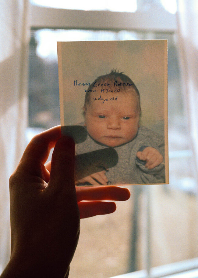 A hand holding a photograph of a newborn baby up to a bight window. At once the head and shoulders image of the baby wearing a blue onesie against a blue and purple backdrop and the inscription on the back can be seen. The hand written inscription reads 'Kenna Grace Robinson born 19 Jan 02 2 days old.