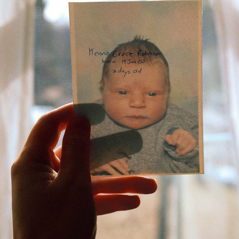 A hand holding a photograph of a newborn baby up to a bight window. At once the head and shoulders image of the baby wearing a blue onesie against a blue and purple backdrop and the inscription on the back can be seen. The hand written inscription reads 'Kenna Grace Robinson born 19 Jan 02 2 days old.