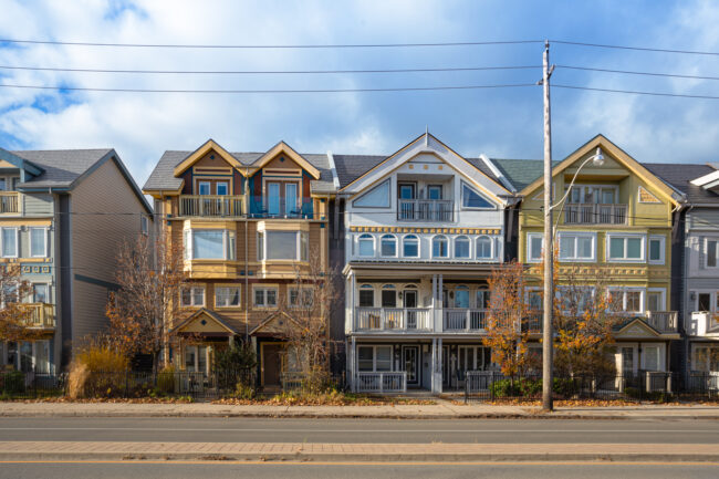 A row of multi-colored townhouses lines a quiet street, displaying a mix of architectural styles and colors. The houses, painted in shades of beige, yellow, gray, and light green, feature unique rooflines, varied window placements, and balconies with decorative railings. Most are two or three stories high, with well-maintained facades. The asphalt-paved street is bordered by a concrete curb, separating it from the sidewalk where bare trees stand, suggesting a late fall setting. The image is taken at eye level, providing a straightforward perspective of the neatly arranged homes.