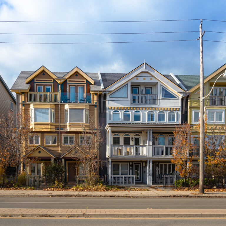A row of multi-colored townhouses lines a quiet street, displaying a mix of architectural styles and colors. The houses, painted in shades of beige, yellow, gray, and light green, feature unique rooflines, varied window placements, and balconies with decorative railings. Most are two or three stories high, with well-maintained facades. The asphalt-paved street is bordered by a concrete curb, separating it from the sidewalk where bare trees stand, suggesting a late fall setting. The image is taken at eye level, providing a straightforward perspective of the neatly arranged homes.