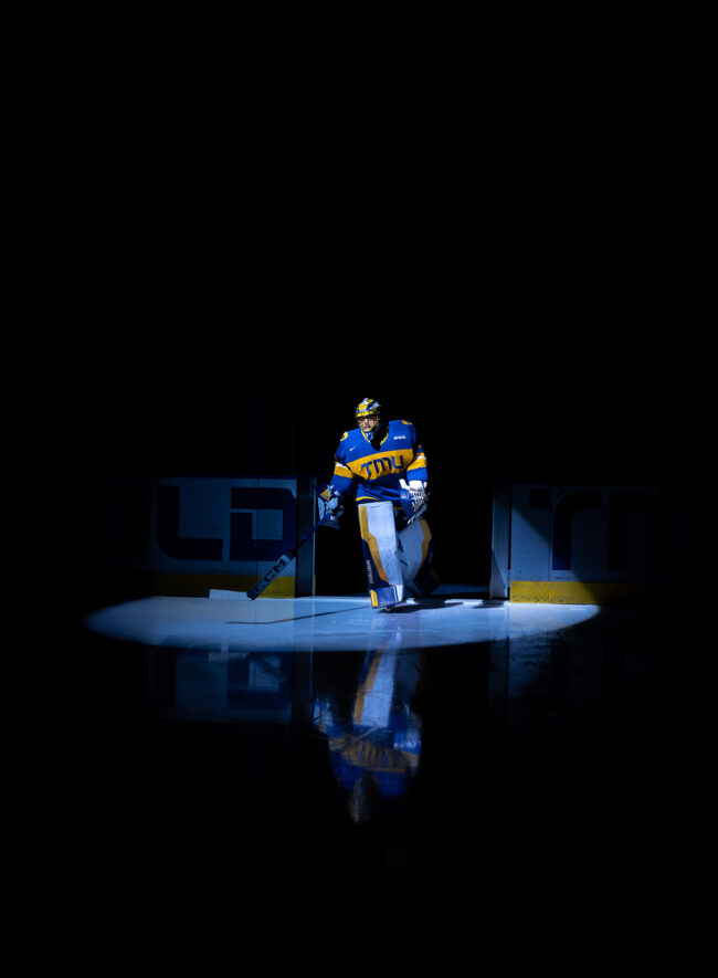 A dramatic shot of a TMU Mens hockey goaltender, Kai Edmonds, stepping onto the ice under a single spotlight.