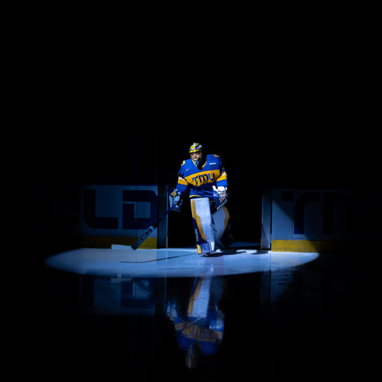 A dramatic shot of a TMU Mens hockey goaltender, Kai Edmonds, stepping onto the ice under a single spotlight.