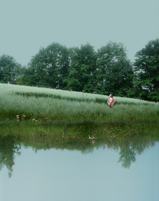 A man wearing a red backpack stands shirtless in a green field clearing with big trees behind him. Underneath him is a reflected image of a different green landscape, almost creating the illusion that he is standing in front of a reflective water surface. Small in the reflected field are two figures crouching in the brush.