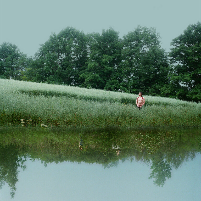 A man wearing a red backpack stands shirtless in a green field clearing with big trees behind him. Underneath him is a reflected image of a different green landscape, almost creating the illusion that he is standing in front of a reflective water surface. Small in the reflected field are two figures crouching in the brush.