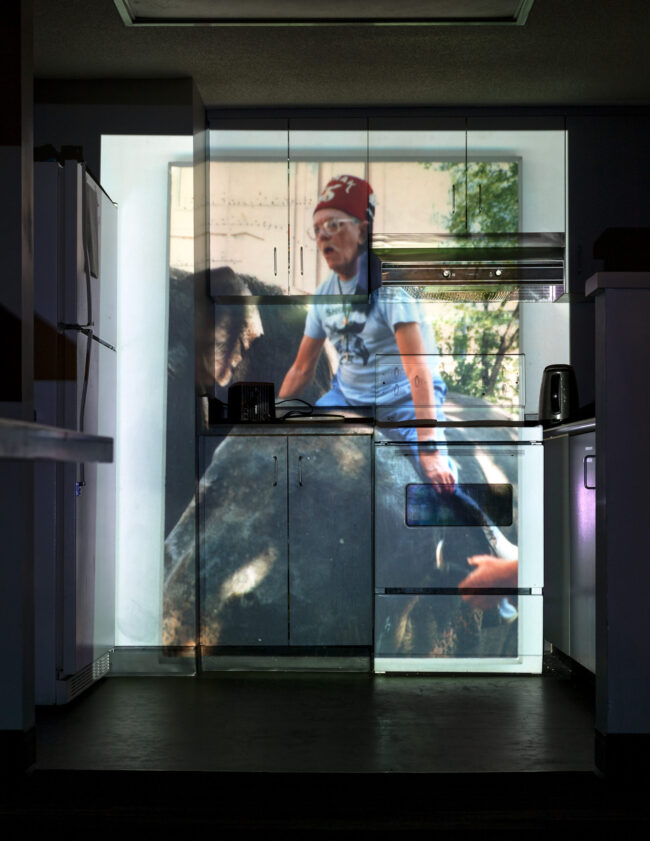 A photograph of a kitchen with a projected image covering the cabinets, oven, and appliances. The projection shows a person wearing a red hat and glasses, sitting on a large animal. The image is aligned with the surfaces of the kitchen, creating a layered visual effect.