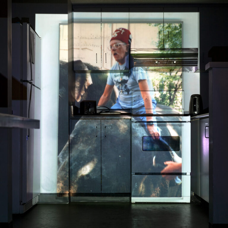 A photograph of a kitchen with a projected image covering the cabinets, oven, and appliances. The projection shows a person wearing a red hat and glasses, sitting on a large animal. The image is aligned with the surfaces of the kitchen, creating a layered visual effect.