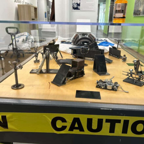 A glass display case showcasing numerous small, dark grey and black metal sculptures, including miniature engines, cranes, and abstract mechanical devices, arranged on a light brown wooden surface. The display case has a black base, around which yellow tape with 'CAUTION' in black letters is prominently wrapped. In the blurred background, a sign reads 'School of Imagine...' suggesting an educational setting, with a hallway, green wall sections, and an orange bottle visible on top of the case.