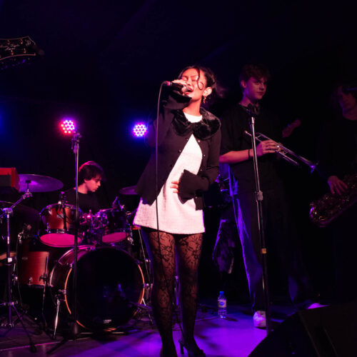A young woman with her eyes closed and mouth open in song stands center stage, holding a microphone. She wears a white mini-dress, a black cardigan with a fuzzy collar, black lace tights, and dark high heels. Behind her, a male drummer plays on the left, and a male trumpeter and saxophonist are visible on the right. The stage is dimly lit by vibrant pink and purple stage lights, highlighting the performers against the dark background.