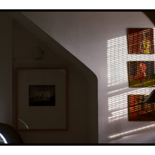 A high-contrast photograph of a room, illuminated by harsh sunlight from the right that projects venetian blind patterns onto a light-colored wall. Three vivid, framed artworks, possibly figurative, in shades of red, yellow, and green are arranged vertically, crisscrossed by the shadows. A dark floor lamp is visible at the bottom right. The left side of the room is shrouded in deep shadow, revealing an angled wall, a large, dark framed print, and a quilted furniture piece.
