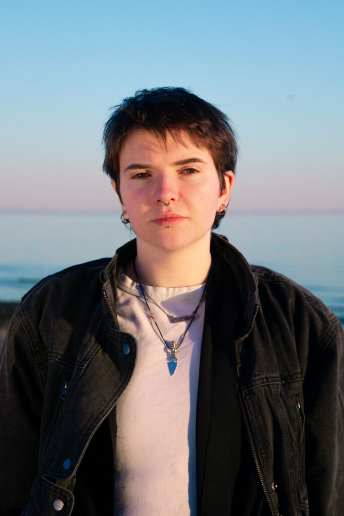 A head-and-shoulders portrait of a light-skinned person with short, dark, slightly messy hair, looking directly into the camera with a neutral expression. They have a silver septum ring, a philtrum piercing, and a labret piercing, along with several silver hoop earrings. They are dressed in a dark, possibly denim, jacket over a white or light-colored t-shirt. Two necklaces are visible: one with a silver dagger-like pendant and another with a silver nameplate. The background is a soft-focus landscape featuring a calm body of water under a clear sky that transitions from light blue to a pale, warm hue at the horizon, suggesting dawn or dusk.