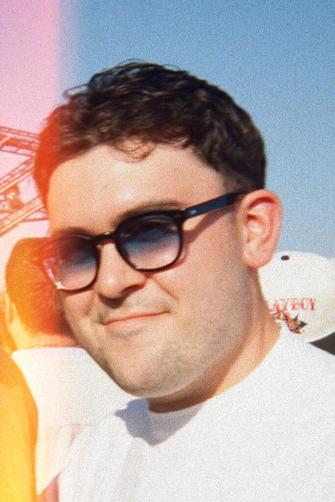 A close-up shot of a fair-skinned man with dark, wavy hair and a slight smile, wearing dark Wayfarer-style sunglasses with blue-tinted lenses and a white polo shirt. The image features a strong film grain texture and a pinkish-orange light leak on the left. The background is blurred, showing a clear blue sky, part of a metal structure like a Ferris wheel on the left, and a white cap with a logo (partially visible 'PLAYBOY') on the right.