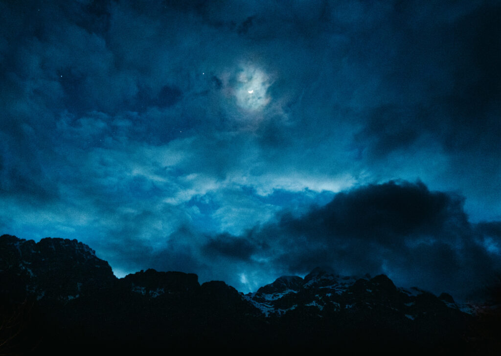 A dramatic long exposure photograph of a deep blue and teal night sky filled with textured, swirling clouds. A luminous crescent moon is visible in the upper-middle right through a break in the clouds, casting a soft glow. Below, the rugged peaks of a mountain range are silhouetted in dark tones against the lighter sky, with a few faint stars scattered among the clouds. The image has a grainy texture, enhancing its atmospheric quality.