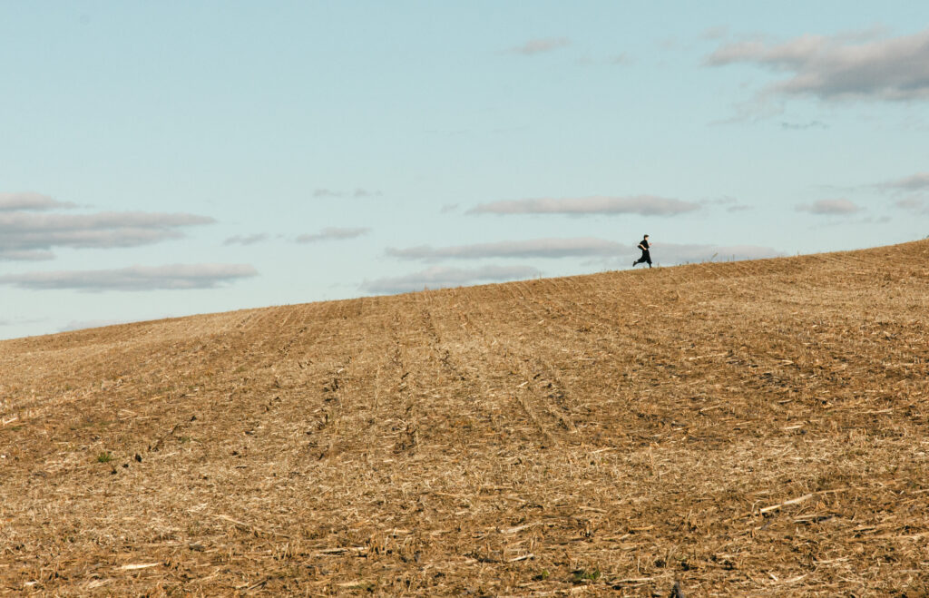 A wide shot shows a dark silhouette of a person running uphill from right to left across an expansive, gently sloping field. The ground is covered in short, golden-brown stubble from a harvested crop, with subtle rows visible. The upper third of the image features a pale blue sky dotted with several thin, light gray clouds, conveying a sense of openness and solitude.