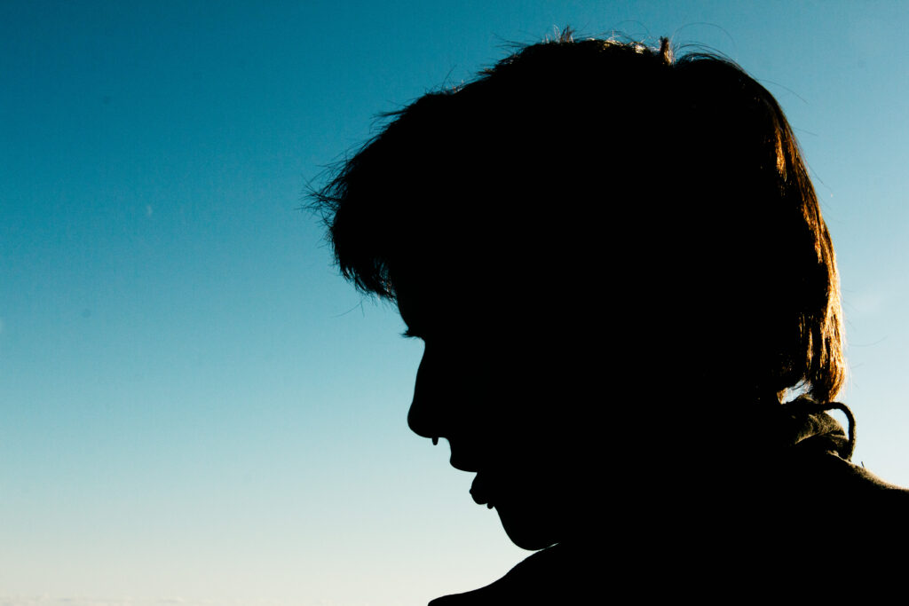 A strong silhouette of a person's head and upper shoulders, seen in profile looking slightly downwards, cast in deep black against a clear blue sky. Bright golden sunlight creates a dramatic rim-light around the person's hair and the outline of their head and shoulder.