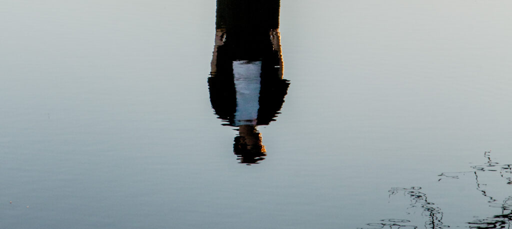 A tranquil scene showing the inverted reflection of a person wearing a dark academic gown with a visible white stripe and a cap in smooth, light gray-blue water. The reflected head at the bottom is softly illuminated by warm, golden light, and subtle ripples create a textured, abstract effect across the water surface, particularly in the lower right.