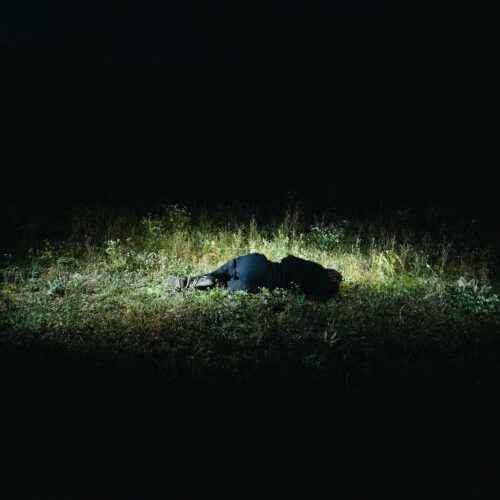 A dramatic, high-contrast image shows a person lying curled up, face down, on a grassy patch. The figure, dressed in dark clothes, is brightly lit by a circular beam of light, while the rest of the frame is enveloped in deep, impenetrable darkness, creating a somber and isolated mood.