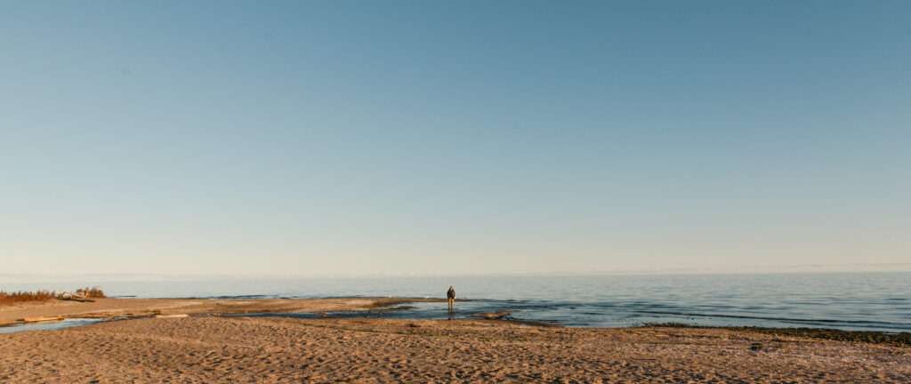 A wide shot of a peaceful sandy beach under a light blue sky. In the midground, a lone person in dark clothing stands in shallow water where a small stream empties into a vast, calm body of water. The foreground features textured light brown sand with some debris and traces of dry grass on the left. The horizon line is flat and clear, separating the tranquil blue water from the pale blue sky.