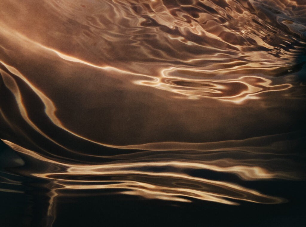 A captivating abstract image showing intricate patterns created by warm golden light refracting and reflecting off the rippling surface of dark water. Luminous, organic curves and streaks of light, appearing like molten gold, dance across the deep brown and black depths, creating a fluid, ethereal visual effect.